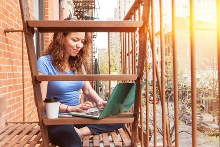 young woman working on her laptop outdoors on a city fire escape just outside her apartment bedroom window