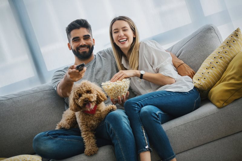 A man, woman and their golden doodle dog snuggle on the couch while watching TV