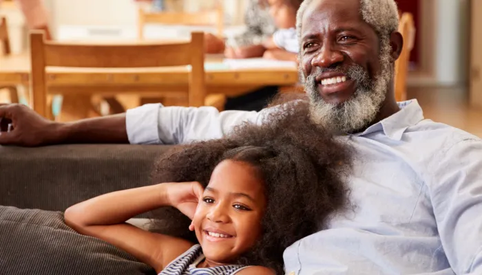 A grandfather and his young grandaughter smile and cuddle on the couch watching TV
