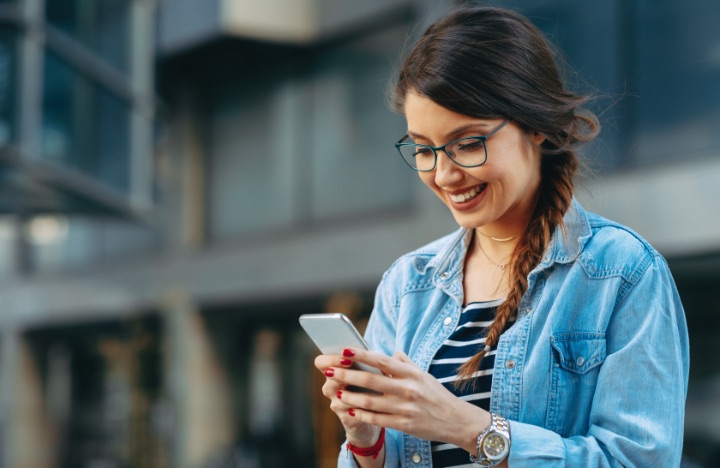 A woman wearing glasses smiles down while texting outside an office building