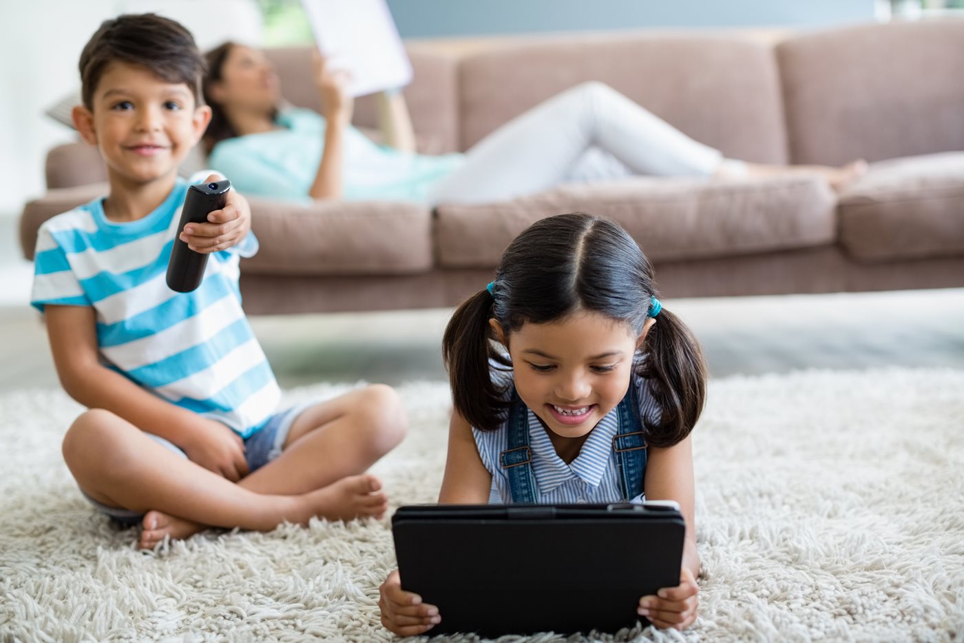 A young boy points a remote at the TV while his sister plays on her iPad