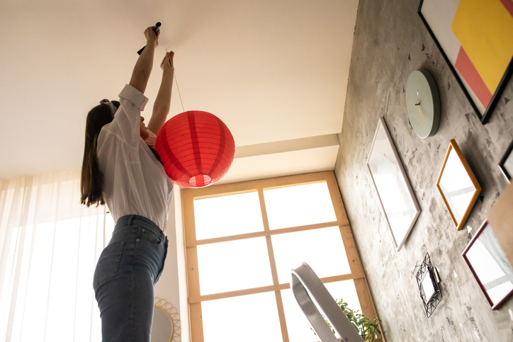 iStock-1253642610.jpg Young woman putting an electric lamp on her high ceilings
