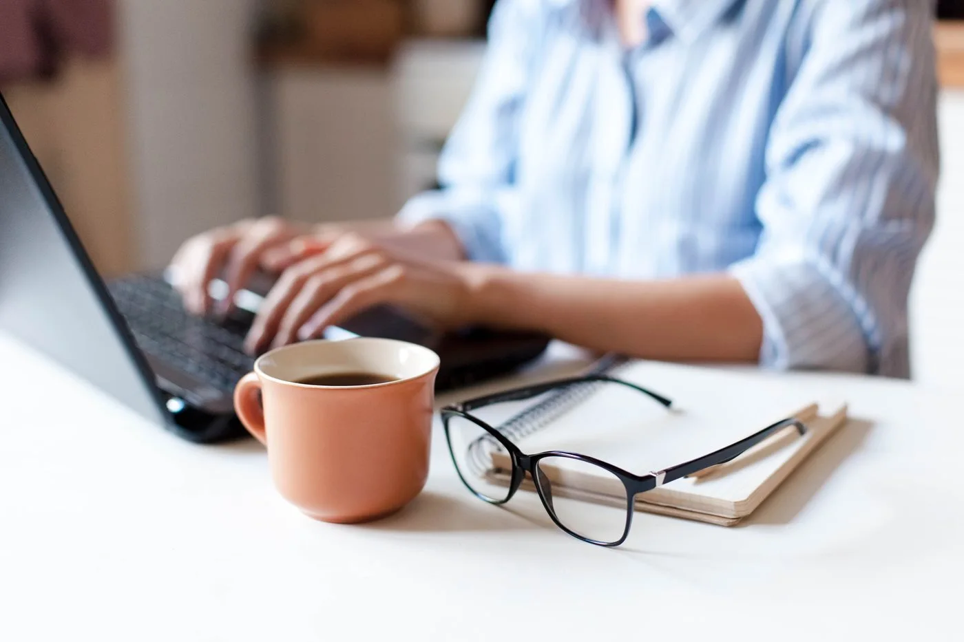 A person's hands typing on a laptop beside a cup of coffee, glasses, and a notebook
