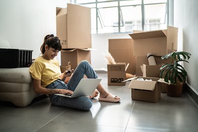 A woman sits on the floor of her new home surrounded by moving boxes while setting up new Internet and TV on her laptop.