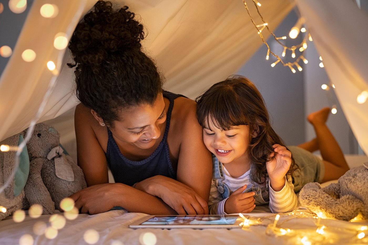 A mom and daughter play on an iPad under a blanket fort with string lights