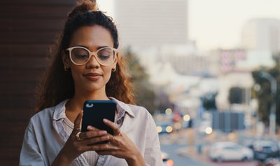 Young woman with glasses looks at her cell phone while walking around a city and getting great coverage and service with a cheap mobile plan.