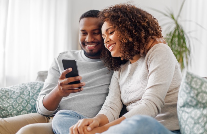 A young couple sit on the couch while smiling at a smartphone