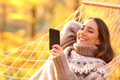 A woman relaxing on a hammock with fall leaves behind her smiles at her phone while streaming content with 3G mobile.