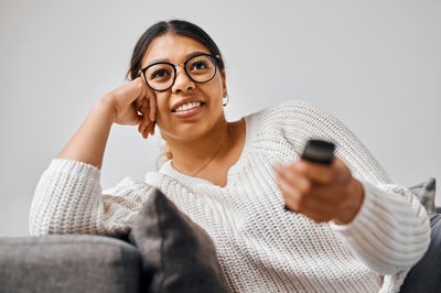 Woman in white sweater and glasses points her remote control at the screen while searching where to watch Emmy Award Winning TV shows.