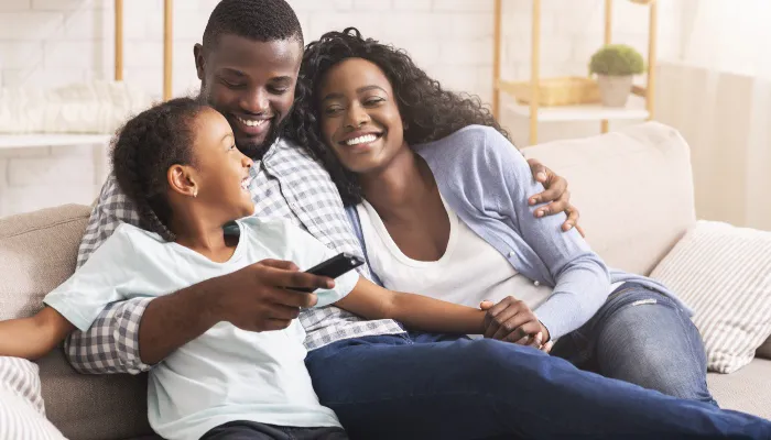 A young daughter happily snuggles on the couch with her mom and dad to watch TV