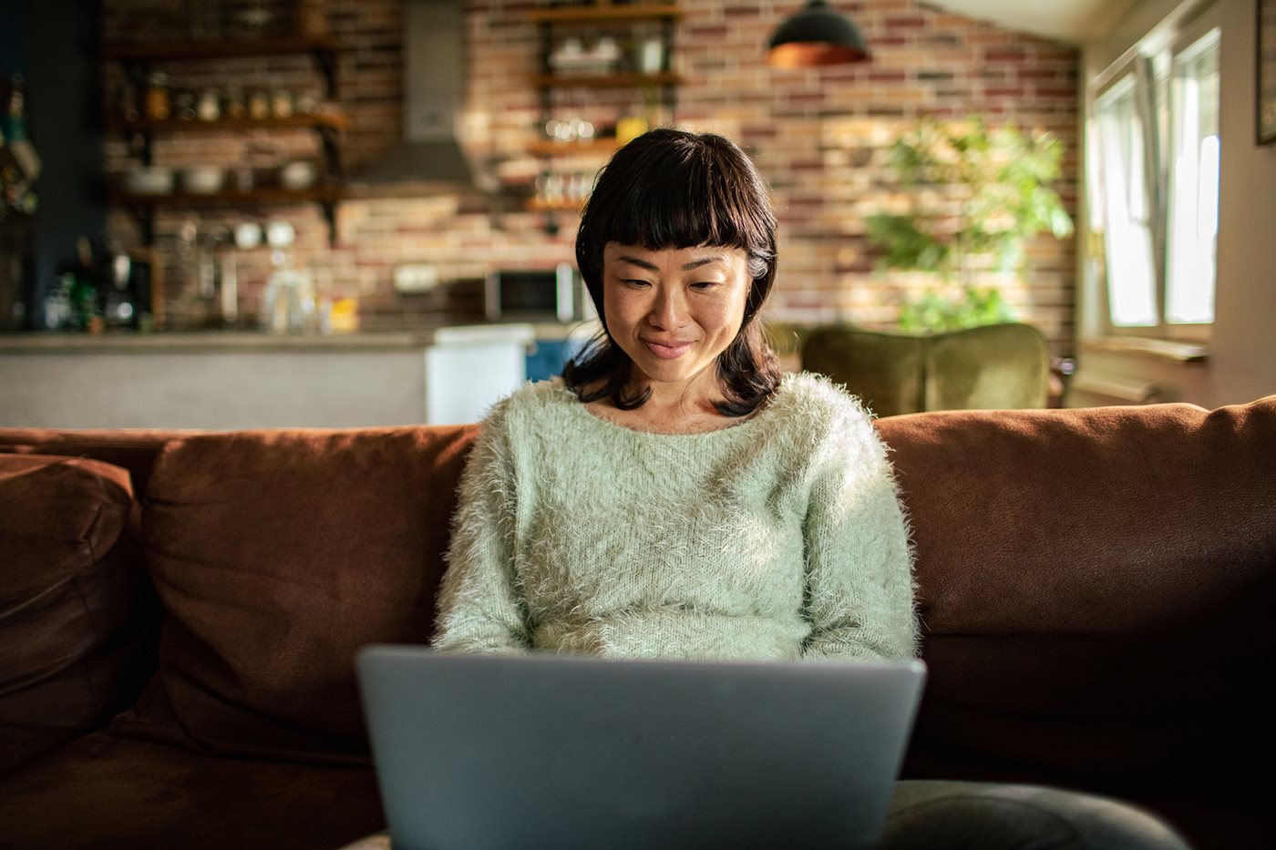 A woman in a sweater sits on her couch while on her laptop exploring GCI Internet, TV and phone plans