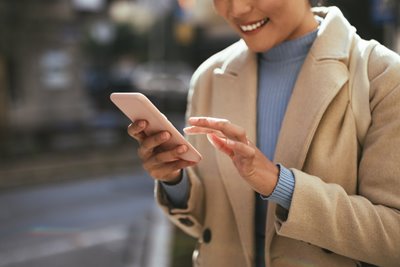A woman in a tan jacket and blue sweater uses her phone with Ting Mobile while walking down a city street.