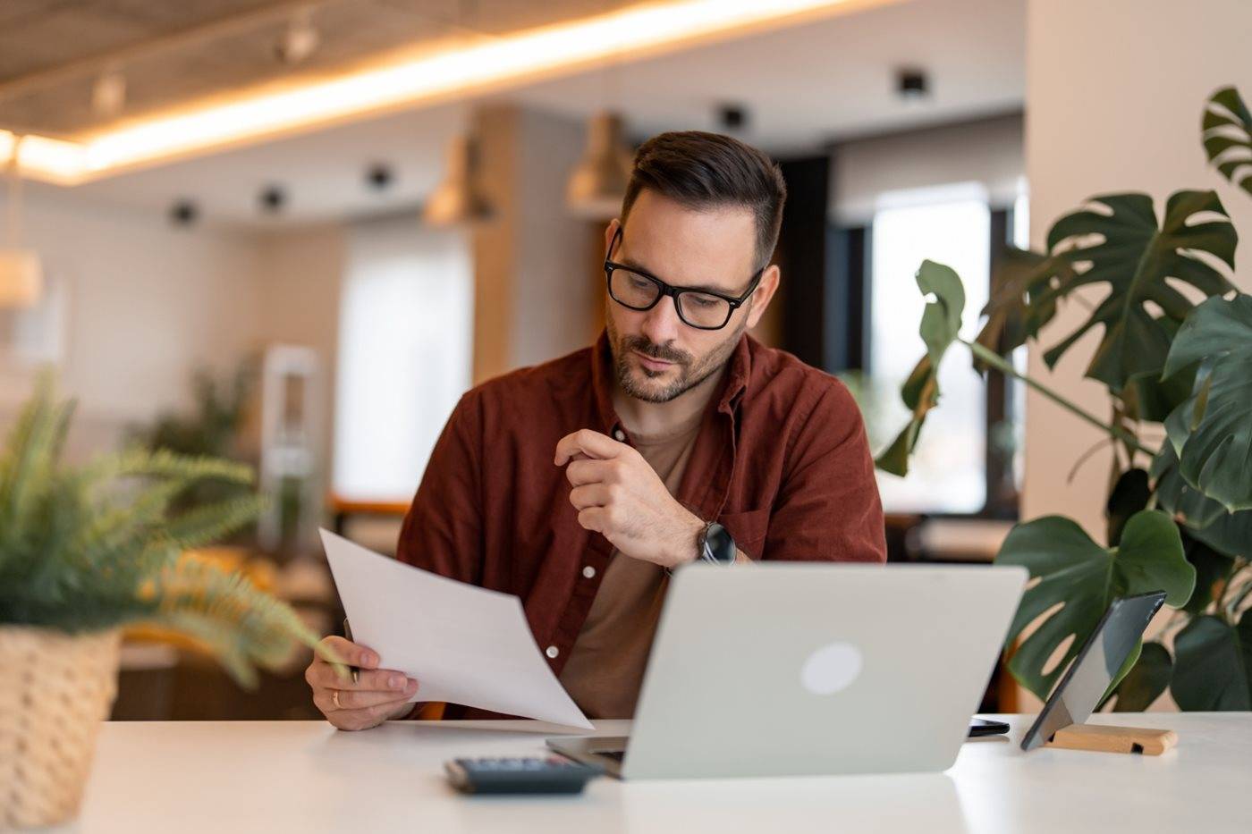 A businessman wearing glasses reviews a paper document while sitting at a desk in front of his laptop.