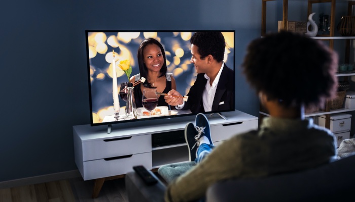 A person faces a television displaying a couple having dinner