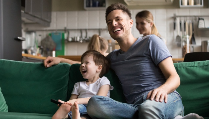 A young father and son laugh on a couch while a mother and daughter are in the kitchen behind them