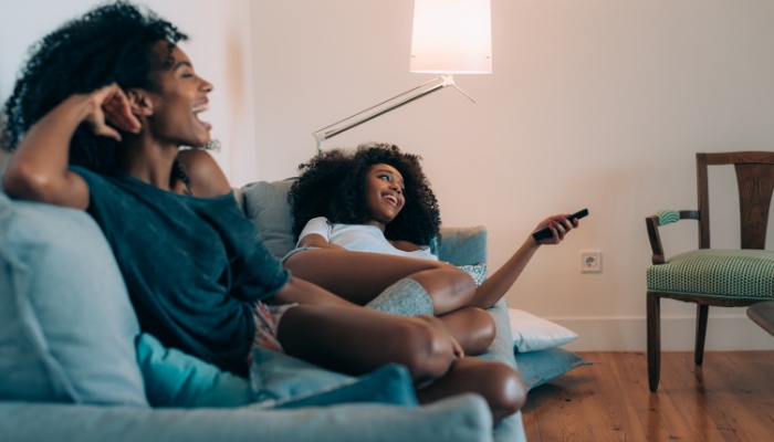 Two young women laugh while sitting on a couch watching TV