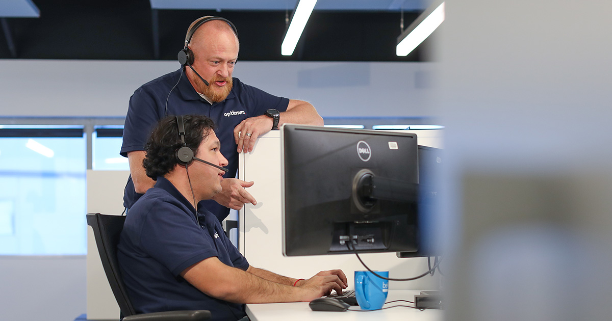 A man works at his computer while another leans over him to help