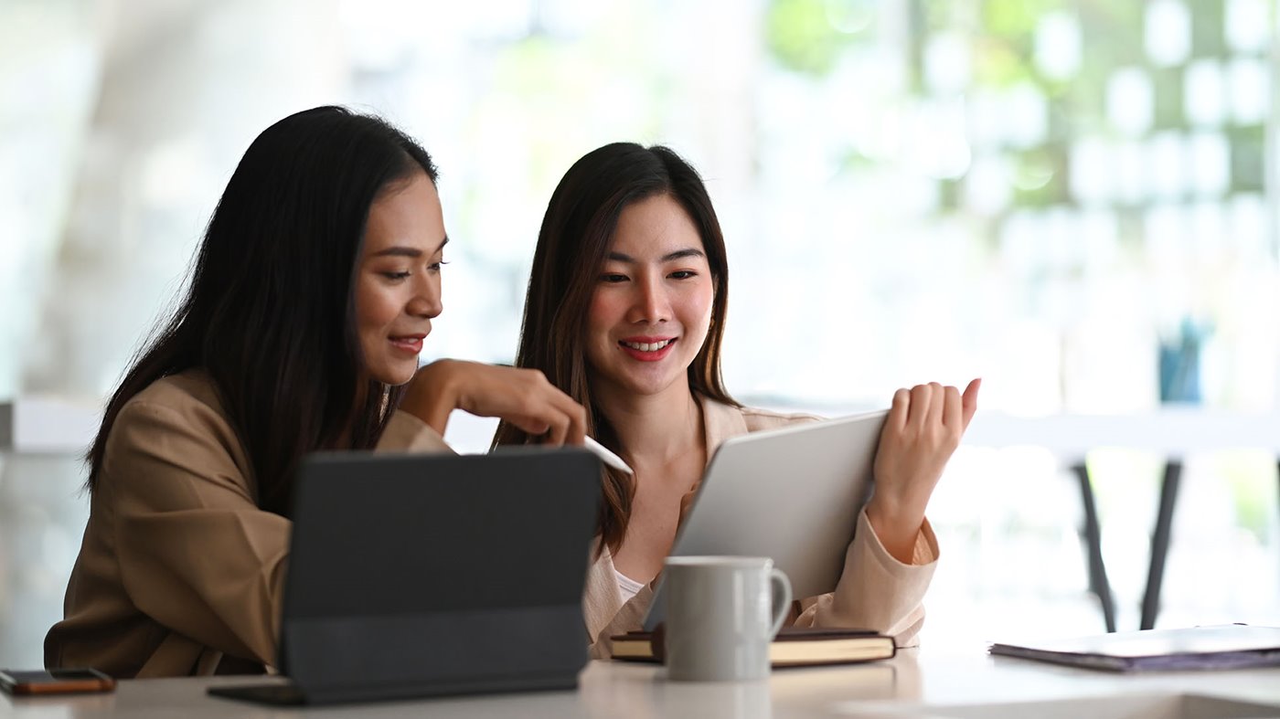 Two young women work together on an iPad and laptop