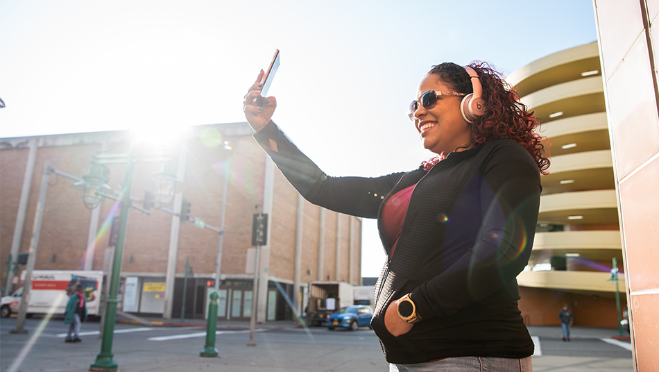 A woman wearing headphones smiles while taking a selfie on her smartphone