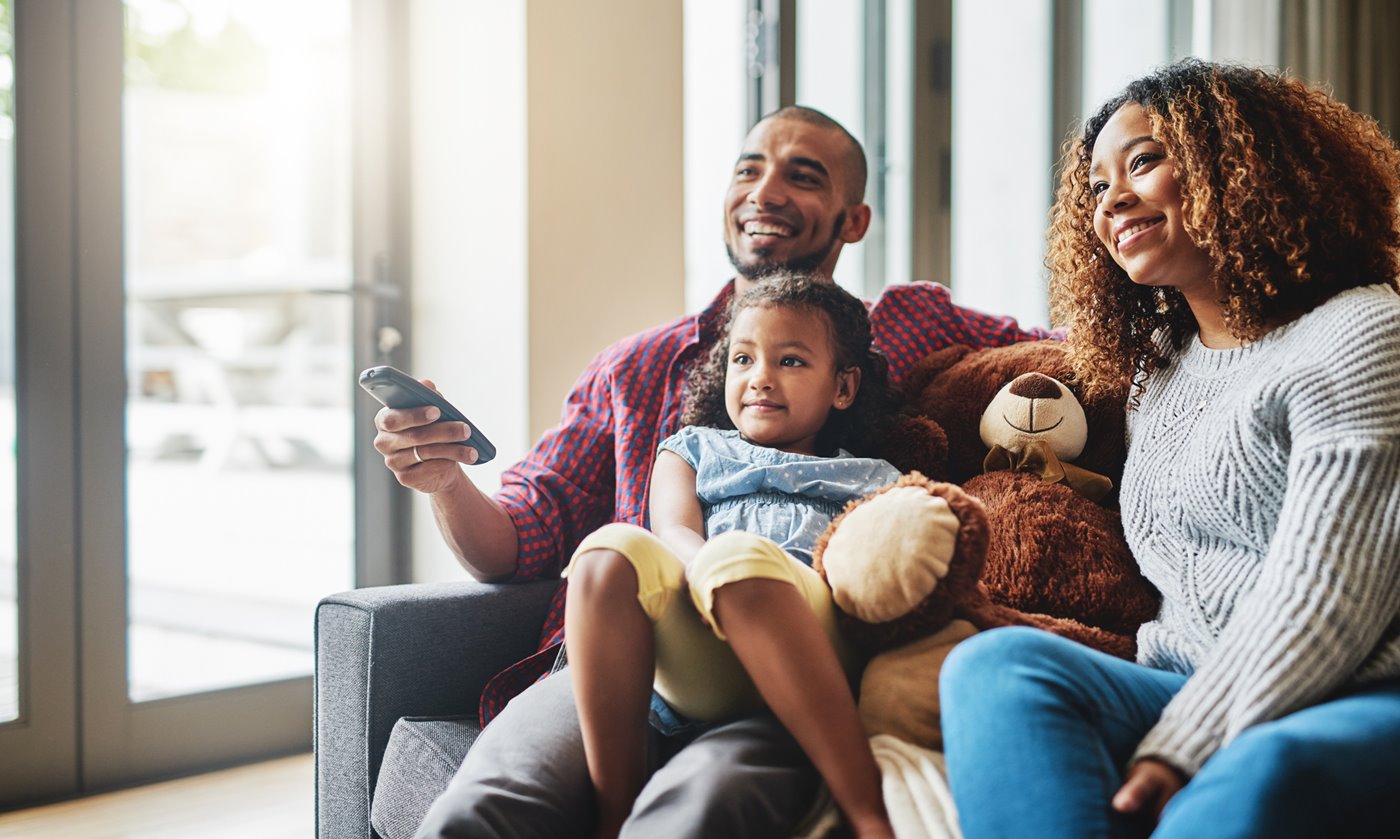 A dad aims a remote at a TV while cuddled up next to his young daughter, her teddy bear, and her mom on the couch