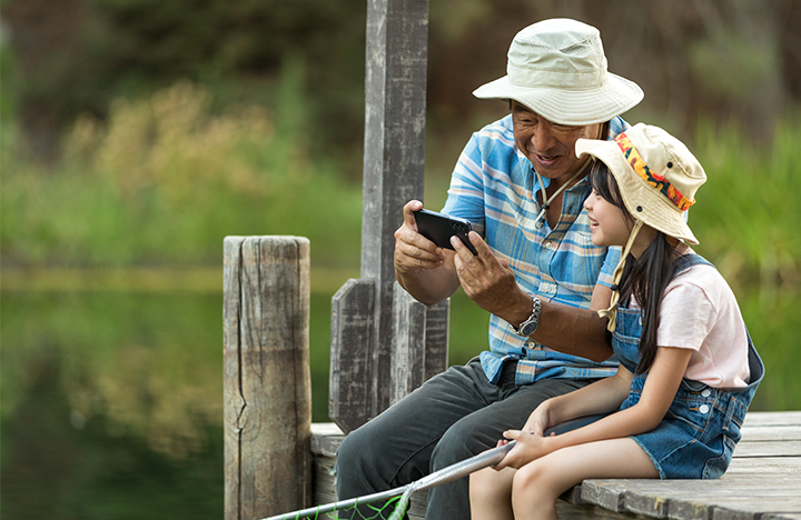 A grandfather and granddaughter sit on a doc wearing fishing hats while smiling at a phone