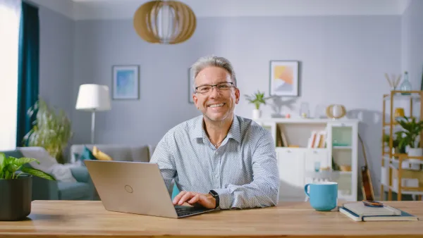An older man smiles up from his desk while typing on his laptop