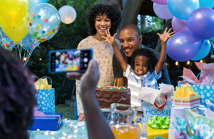 A mom, dad and young daughter pose for a photo while surrounded by balloons and presents on her birthday