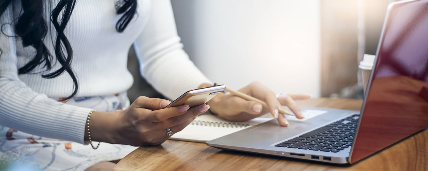 A woman types on a laptop while holding her mobile cell phone in one hand