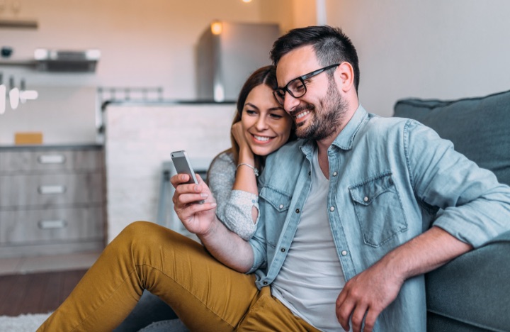 A young couple sit on their living room floor while smiling at the same smartphone