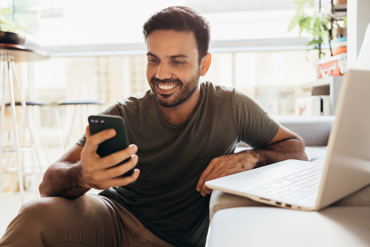 A man smiles at his phone while sitting in front of his laptop