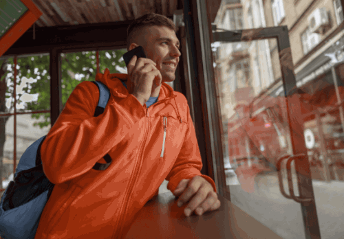 A man with a backpack smiles while holding his smartphone to his ear and looking out a window