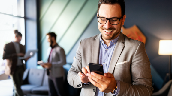 A businessman smiles down at his mobile device while smiling coworkers gather in the office behind him