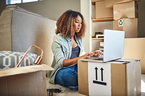 A woman sits on the floor surrounded by moving boxes while using her laptop