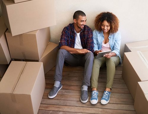 A young couple sits on the floor surrounded by moving boxes while looking at an iPad