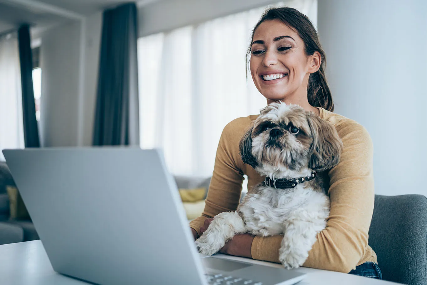 A happy young professional woman holds her dog while working on her laptop