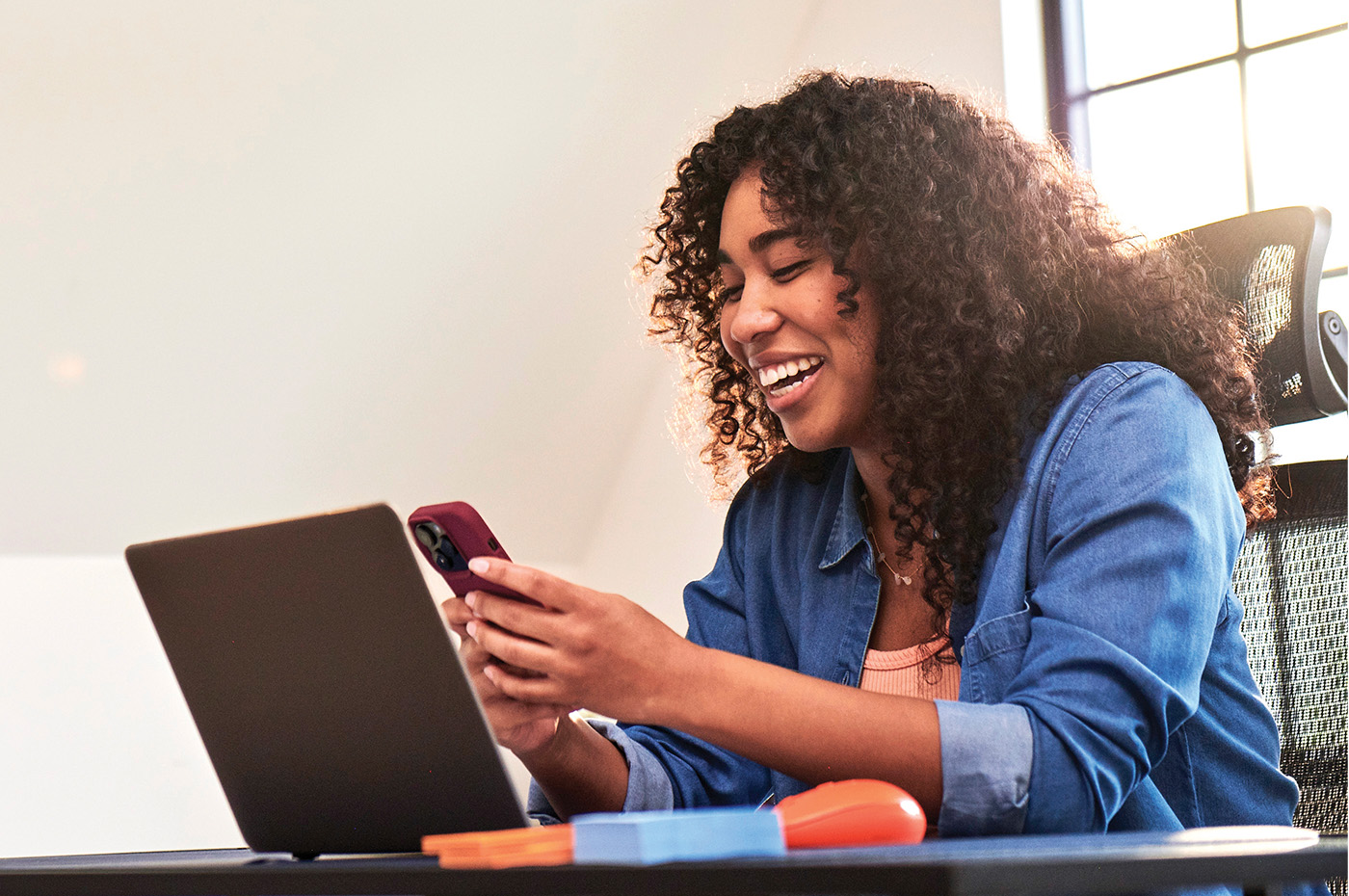 A young woman smiles at her desk while using her phone and laptop powered by Optimum