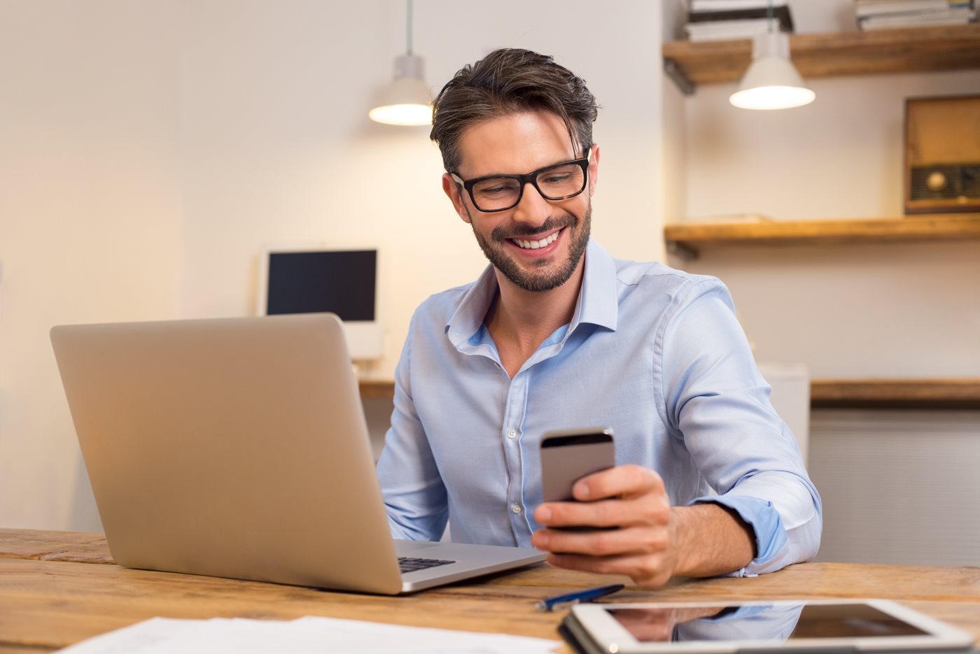 A businessman sits in front of a laptop and smiles while looking at his mobile device