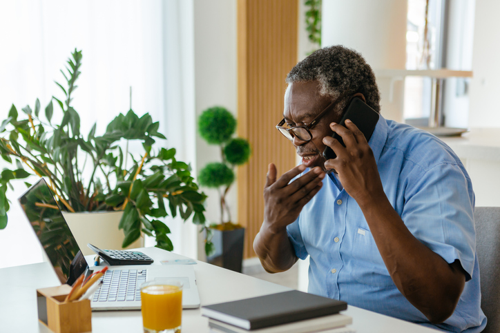 An older man sits at a desk on the phone while looking at his laptop