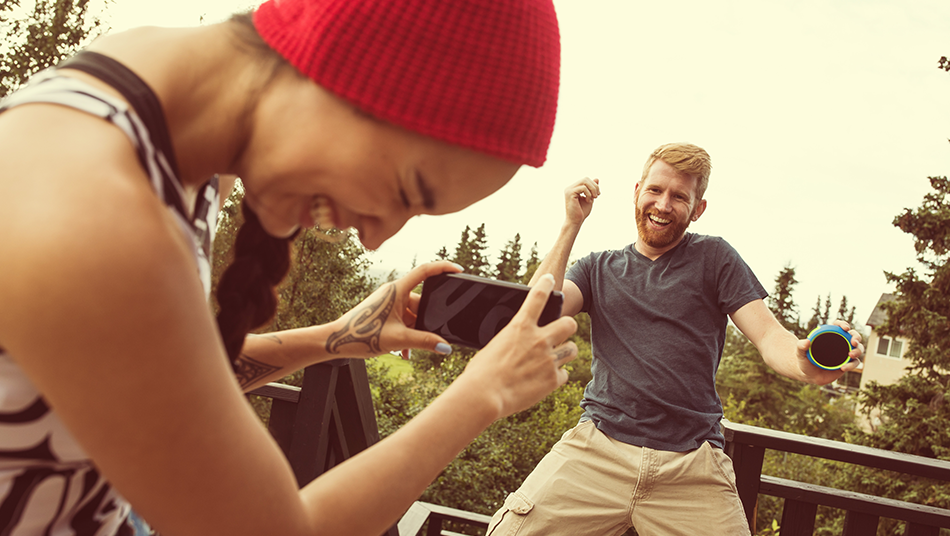 A smiling woman in a beanie takes a photo of her dancing male friend on her smartphone
