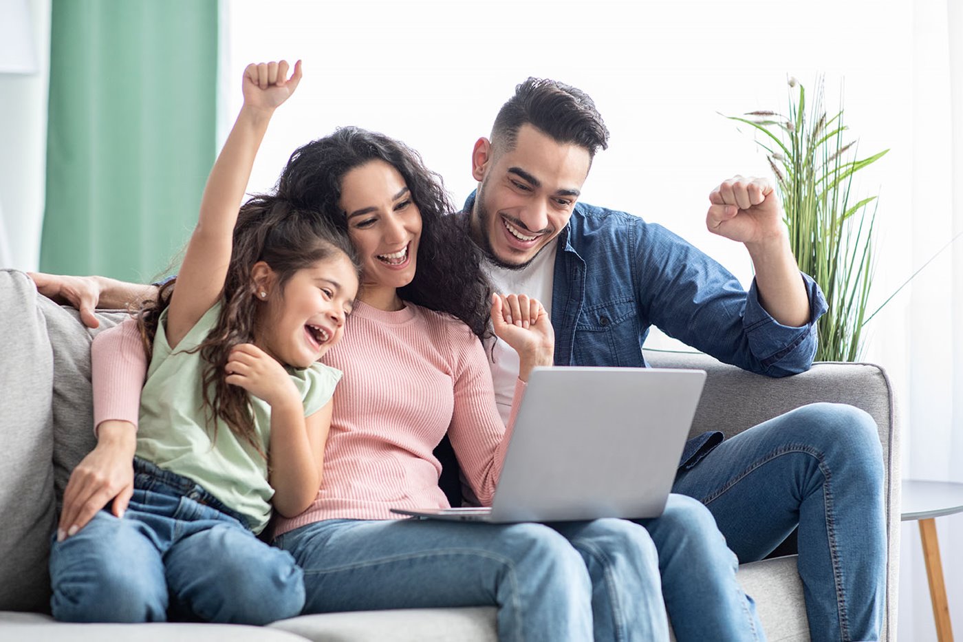 A young family snuggle on the couch while cheering at their laptop