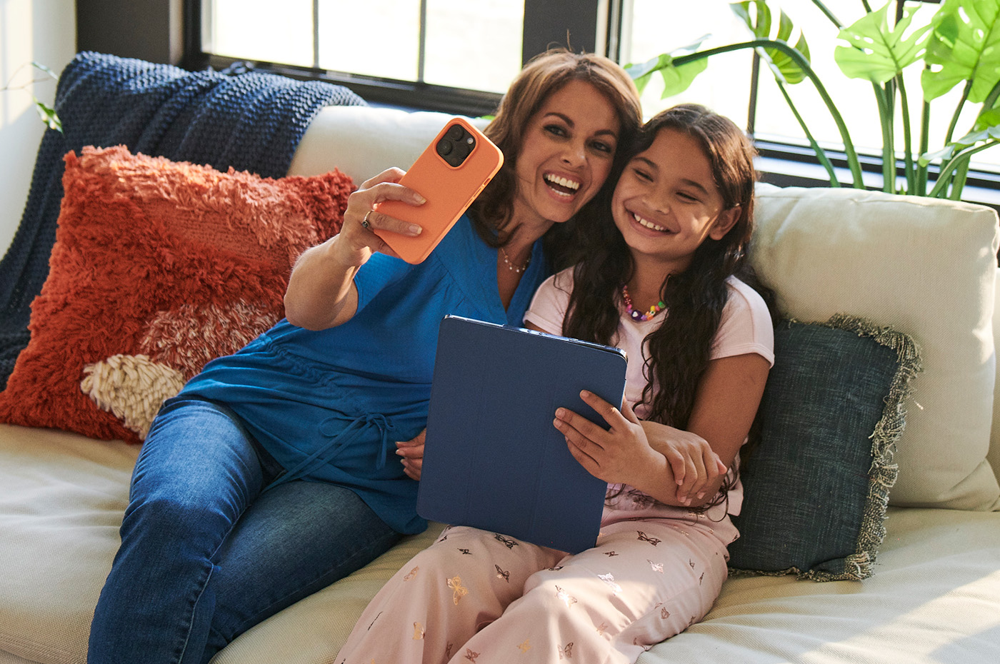 A mother and daughter smile on the couch as one takes a selfie with a phone and the other holds an iPad, both powered by Optimum Internet