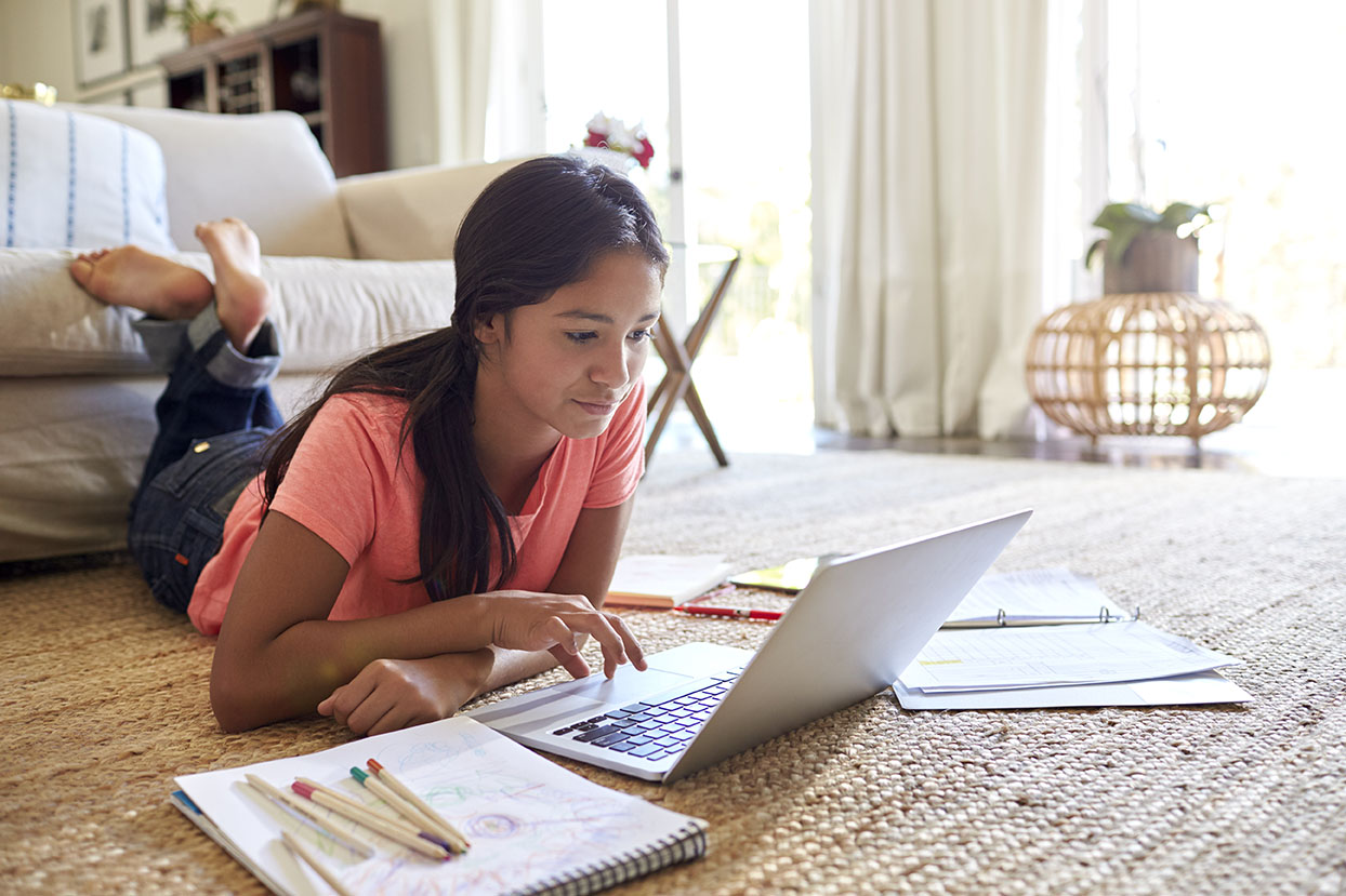 A young girl lays on the floor while doing homework on her laptop