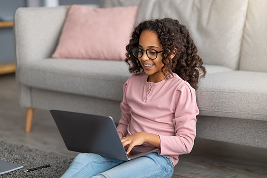 A young girl uses her laptop while sitting on the floor and leaning against a couch