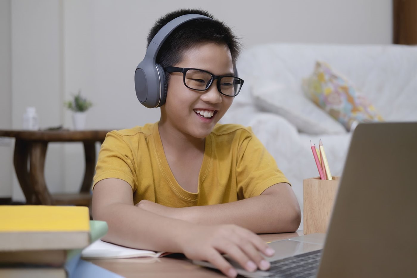 A young boy wears a headset and smiles while doing schoolwork on his laptop
