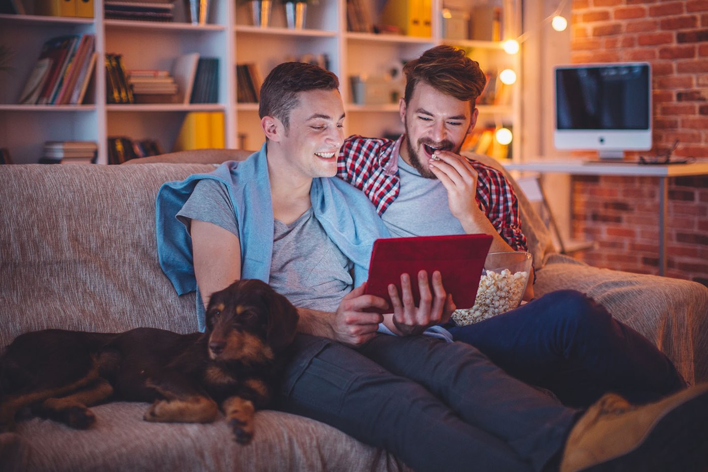 Two men sit on the couch next to their dog while smiling at an iPad and eating popcorn